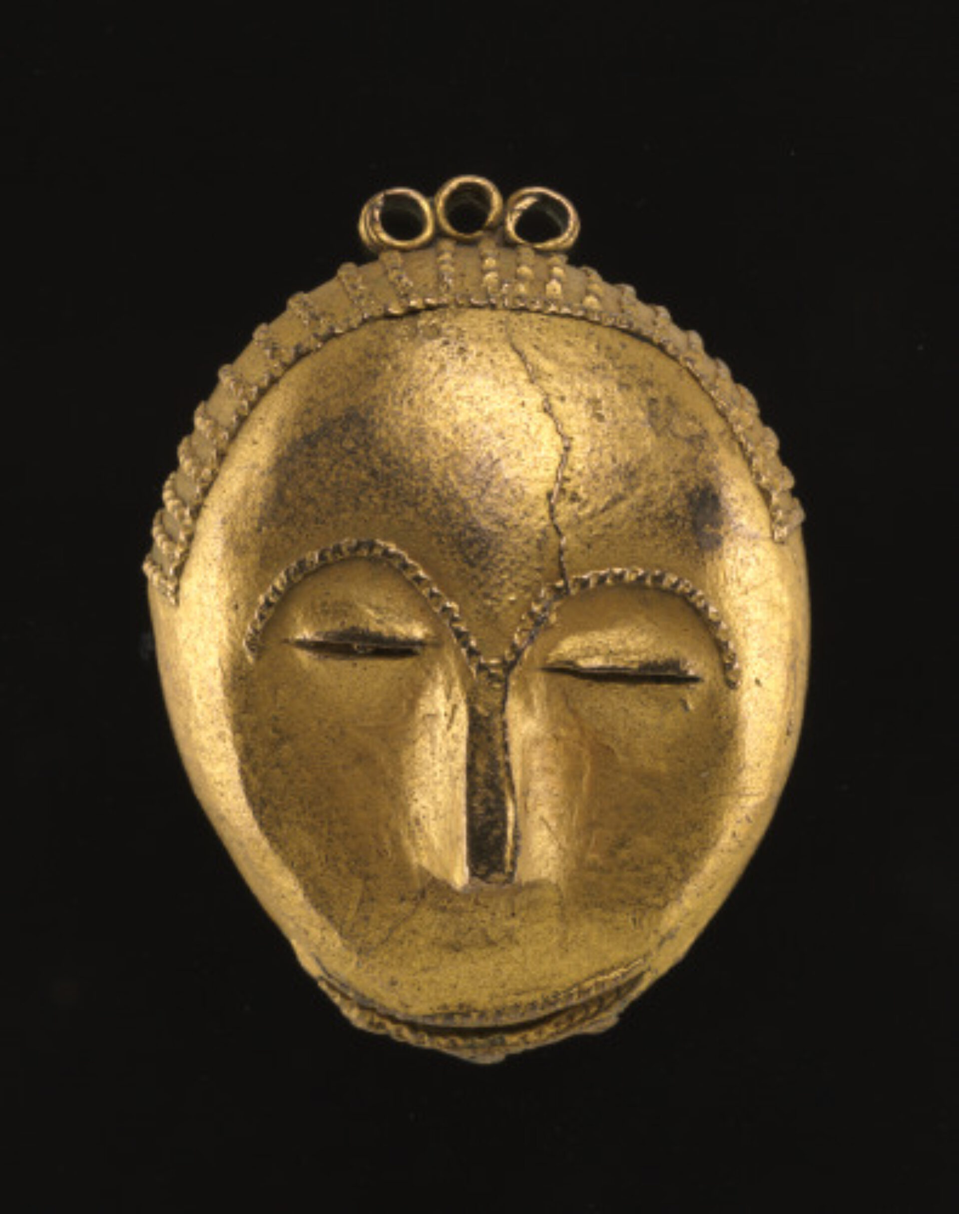 A miniature gold-alloy mask with closed eyes, a narrow nose, and a subtle smile. The top edge of the mask is textured with a pattern resembling braided hair.