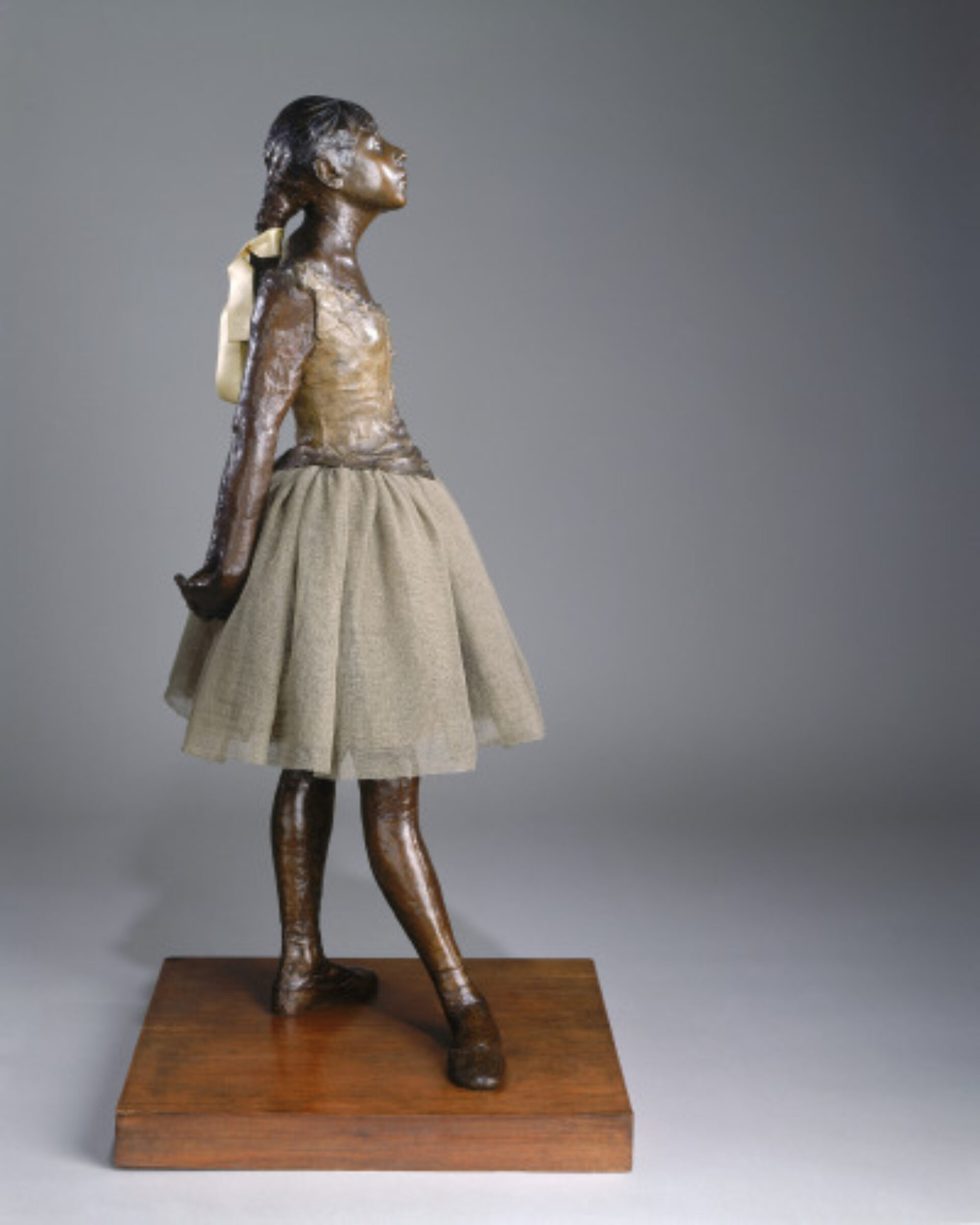 A bronze sculpture of a young ballet dancer appearing as a girl. The figure poses with her head slightly tilted upwards and her arms gently extended behind her. She is wearing a textured, fabric tutu and her hair is styled in a braid tied with a ribbon.